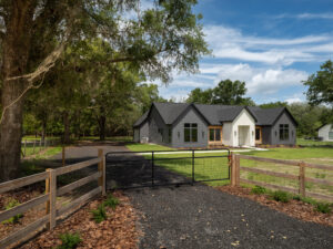A modern single-story house with dark siding and white accents sits behind a wooden fence and metal gate, surrounded by trees and a gravel driveway.