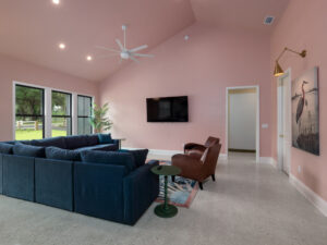 Living room with pink walls, blue sectional sofa, brown armchair, wall-mounted TV, potted plant, and large windows overlooking greenery.