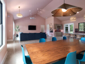 Modern open-plan living and dining area with pink walls, wood dining table, blue chairs, navy sofa, and large windows providing natural light.
