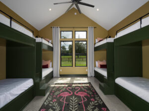 Room with four green built-in bunk beds, white bedding, a flamingo-patterned rug, two white curtains, and a window overlooking a grassy field and trees. Ceiling fan above.