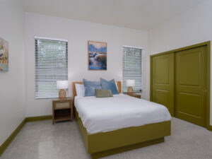 Bedroom with a green bed frame, matching closet doors, white bedding, two nightstands with lamps, and two windows with blinds. A landscape painting hangs above the bed.