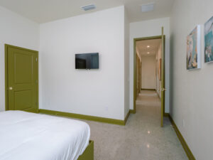 Bedroom with white walls, green trim, a wall-mounted TV, partially visible bed, open door, framed art, and a view of a hallway with wall art and ceiling lights.
