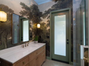 Bathroom with a marble countertop, wooden vanity, wall sconces, large mirror, frosted glass door, and mural of trees on the walls.