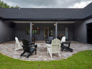 A stone patio with six Adirondack chairs arranged around a circular fire pit, outside a dark gray house with covered porch and glass doors.
