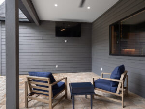 Covered patio with two wooden chairs featuring blue cushions, a small black table between them, and a wall-mounted TV on the gray paneled wall. Brick paver flooring is visible.