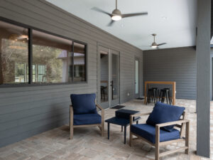 Modern covered patio with two navy cushioned chairs, a small table, a dining table with stools, ceiling fans, brick flooring, and gray siding walls.