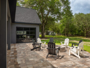 A paved patio with six Adirondack chairs arranged around a fire pit, next to a gray house and surrounded by green trees and grass.
