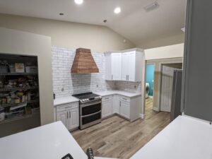 Modern kitchen with white cabinets, a wooden vent hood, tile backsplash, stainless steel stove, open pantry, and light wood floors; a laundry room is visible through an open door.