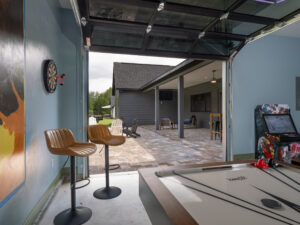 A game room with bar stools, dart board, air hockey table, and arcade machine opens to a covered patio with chairs and a ceiling fan.