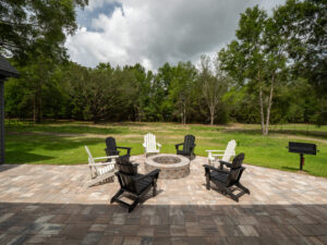 Six Adirondack chairs surround a circular stone fire pit on a paved patio, with a grassy yard, trees, and a barbecue grill in the background under a partly cloudy sky.