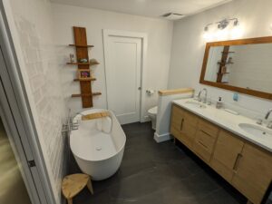 Modern bathroom with a freestanding tub, wooden shelves, double sink vanity with a large mirror, and white walls with dark tile flooring.