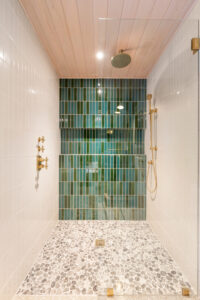 Modern shower with green vertical tile accent wall, white side tiles, pebble stone floor, glass door, and brass fixtures including rain showerhead and handheld sprayer.