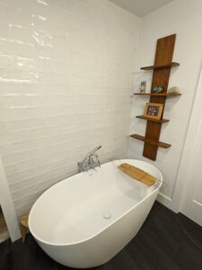 Modern white bathtub with a wooden bath tray, set against a white tiled wall. Wooden corner shelves hold a photo frame, a stone, and a small metal object.