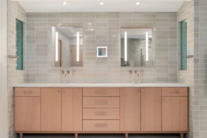 Modern bathroom with double sinks, light wood cabinets, two mirrors with vertical lights, and beige tiled walls.
