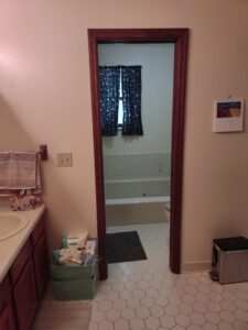 Bathroom with a sink, toiletries bin, towel, and calendar. Through the doorway is a tiled bathtub area with a dark mat and a window with a blue patterned curtain.