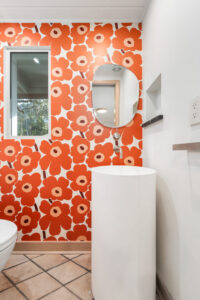 Bathroom with bold red and orange floral wallpaper, a round mirror above a cylindrical white sink, a window, and tiled floor.