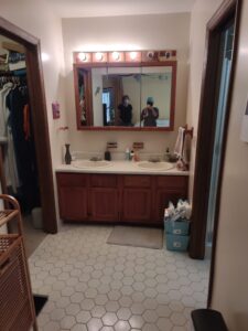 Two people in masks are reflected in a bathroom mirror above a double sink vanity. The bathroom has hexagonal tile flooring and wooden trim, with a closet visible to the left.