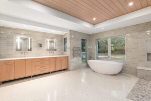 Spacious modern bathroom with wood cabinets, double sinks, large mirrors, a freestanding oval bathtub, beige tiled walls, and large windows overlooking a green outdoor area.