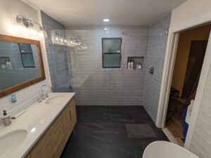 Modern bathroom with dual-sink vanity, large mirror, gray tile walls, rain shower, and black tile floor. Bottles are visible on a window ledge and built-in shelf. Doorway opens to another room.