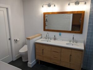 Modern bathroom with dual-sink wooden vanity, large mirror, wall lights, and a toilet near the door; light tile and white walls.