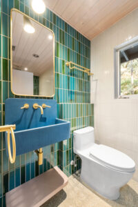 Modern bathroom with green tile wall, blue rectangular sink, gold fixtures, white toilet, large mirror, towel rack, and a window showing trees outside.