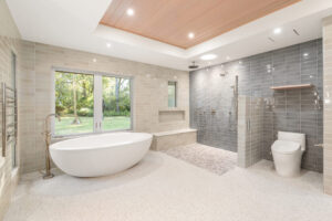 Modern bathroom with freestanding tub, walk-in shower, and toilet. Large windows overlook a green yard. Neutral tiles and wood ceiling design detail.