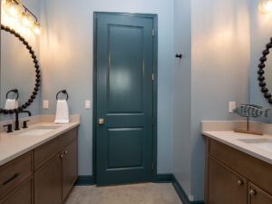 Modern bathroom with blue walls, a central blue door, two vanities with round mirrors, black fixtures, and globe light bulbs above each sink.