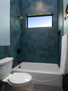 Modern bathroom with teal square tiles, a white toilet, a bathtub with a black showerhead, a window above the tub, a towel rack, and a small wall mirror visible on the right.