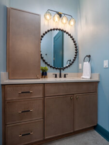 Modern bathroom vanity with light wood cabinets, round beaded mirror, black fixtures, three-bulb light fixture, and a small potted plant on the countertop.