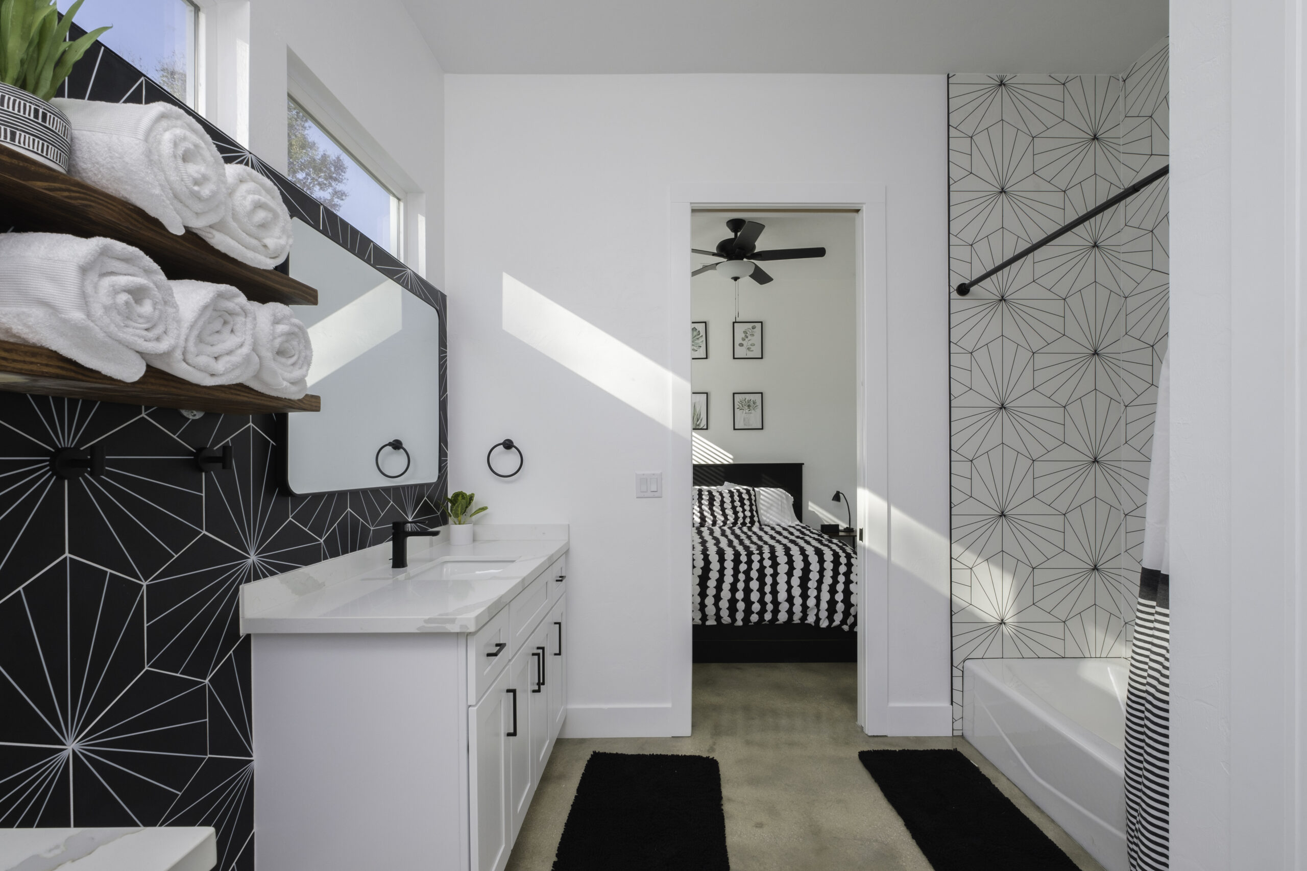 Modern bathroom with geometric black-and-white tile, double vanity, open shelving with towels, and view into a bedroom with matching decor.