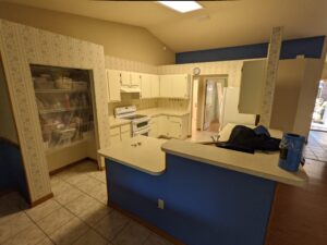 A kitchen with cream cabinets, patterned wallpaper, white appliances, tiled floor, and a blue accent wall. The counter holds a few items, including a rolled-up towel and a canister.