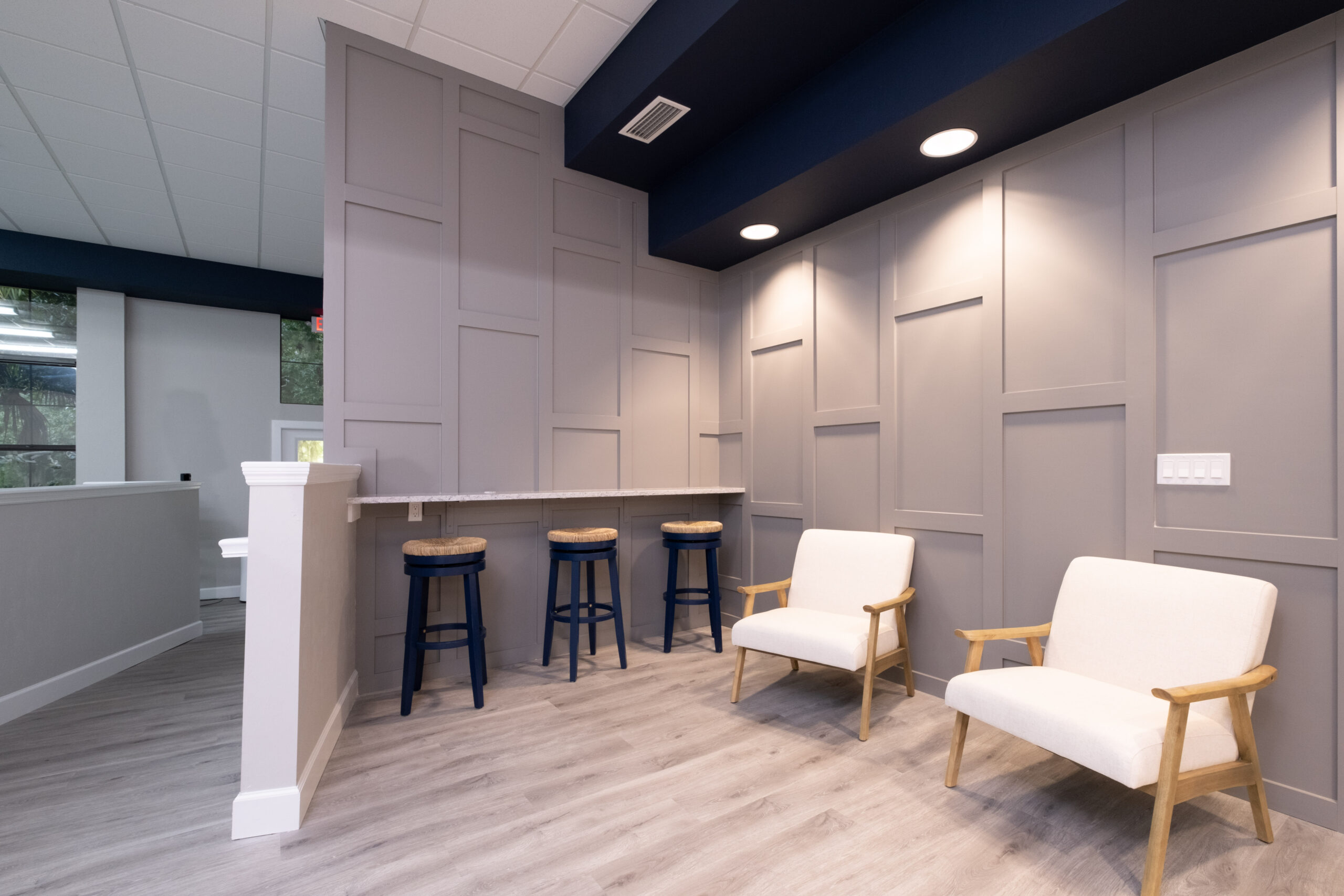 A modern waiting area, perfect for home remodeling inspiration, features two white armchairs, a counter with three blue stools, paneled gray walls, and wood flooring.