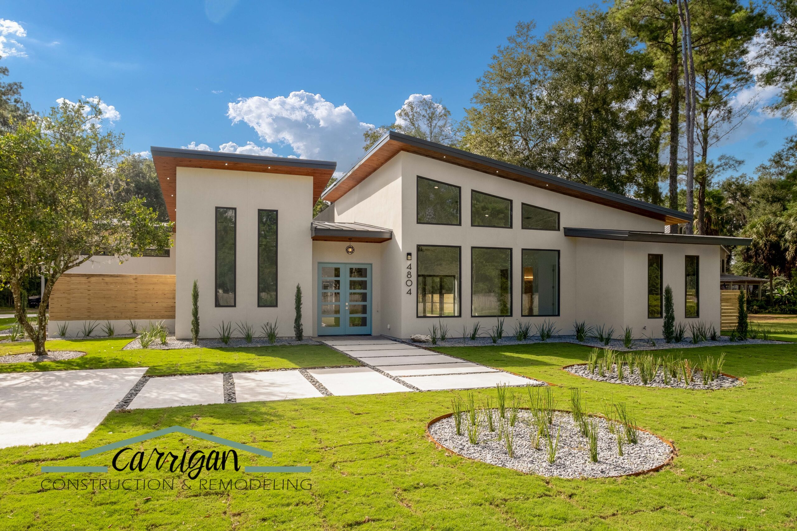Modern single-story house with large windows, sloped roof, and a landscaped front yard; “Carrigan Construction & Remodeling” logo in the lower left corner.