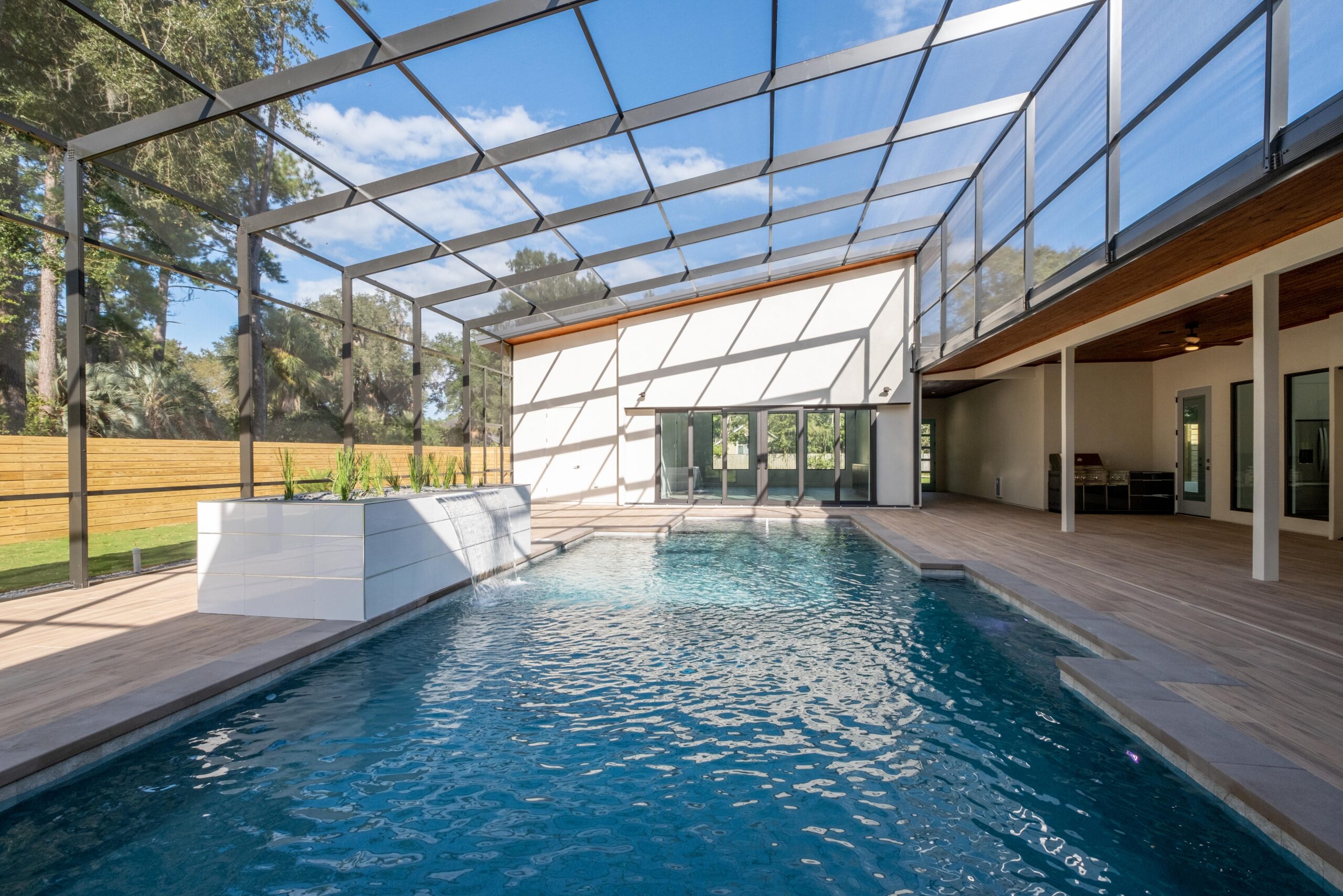 Indoor-outdoor pool area with a screened enclosure, wooden deck flooring, and large windows, under a clear blue sky.