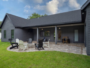 A modern gray house with a covered patio, Adirondack chairs around a fire pit, and a paved seating area on a grassy lawn under a partly cloudy sky.