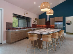 Modern kitchen with a large marble island, six wicker barstools, wood cabinets, red tile backsplash, pendant lights, and a teal accent wall.