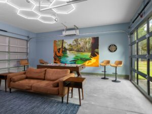 A modern game room with a tan sofa, pool table, barstools, dartboard, large wall art, glass garage doors, and hexagonal ceiling lights.