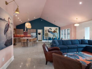Open living space with blue sectional, brown leather chair, kitchen with bar stools, pendant lights, and large bird artwork on pink walls.