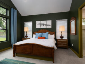 Bedroom with dark green walls, wooden bed with white bedding, blue pillows, two bedside tables with lamps, and three-panel whale artwork above the headboard.