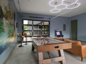 Game room with an air hockey table, brown sofa, bar stools, a wall mural, arcade machine, and a geometric light fixture. Glass garage door leads to an outdoor area.