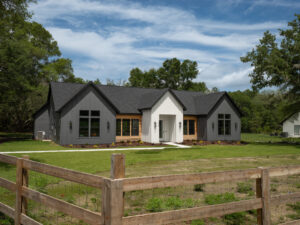 Modern farmhouse with dark gray and white exterior, large windows, and double front doors, set behind a wooden fence with a grassy yard and trees in the background under a partly cloudy sky.