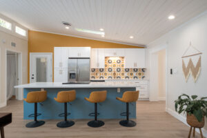 Modern kitchen with white cabinets, a patterned tile backsplash, stainless steel appliances, a blue island, and four yellow barstools. Wood flooring and a plant are also visible.