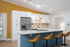 Modern kitchen with a yellow accent wall, patterned backsplash, white cabinets, stainless steel appliances, blue island, and four tan bar stools.