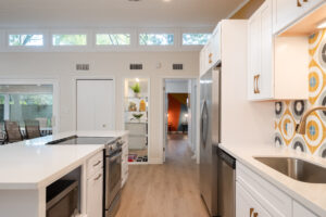Modern kitchen with white cabinets, stainless steel appliances, patterned backsplash, and a view into a brightly lit living space with orange accent wall and decorative shelves.