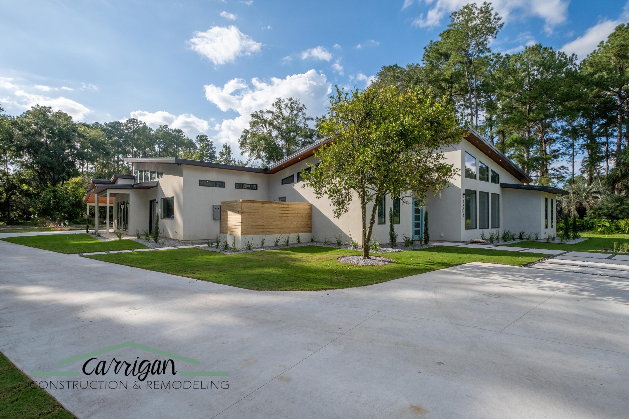Modern, single-story home with large windows, angular rooflines, light exterior, and landscaped yard surrounded by trees. Carrigan Construction & Remodeling logo at the bottom left.