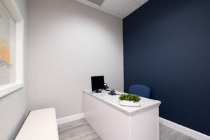 Small modern office with a white desk, computer monitor, telephone, potted plant, blue office chair, and a dark blue accent wall. A bench sits against the opposite wall.