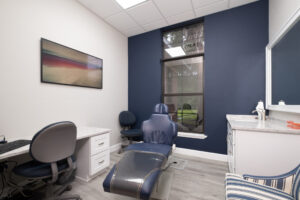 A modern dental exam room with a reclining chair, computer station, cabinets, large window, and blue accent wall.