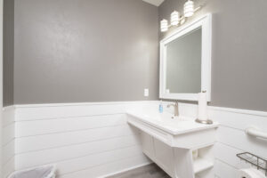 A modern bathroom with gray and white walls, a rectangular mirror, overhead lights, a white sink, a soap dispenser, and a paper towel roll.