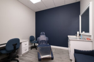 Modern dental exam room with a reclining chair, two desks, office chairs, a sink, a large mirror, and a dark blue accent wall.
