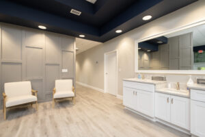 Modern waiting room with two beige chairs, wood flooring, white cabinets with a granite countertop, and a large mirror on the wall under recessed lighting.